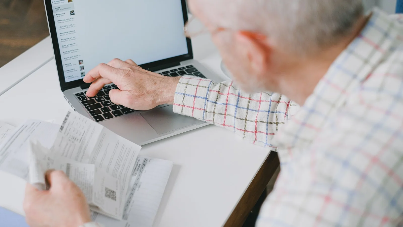 Older man typing on a laptop while holding a stack of printed bills and statements at a white desk