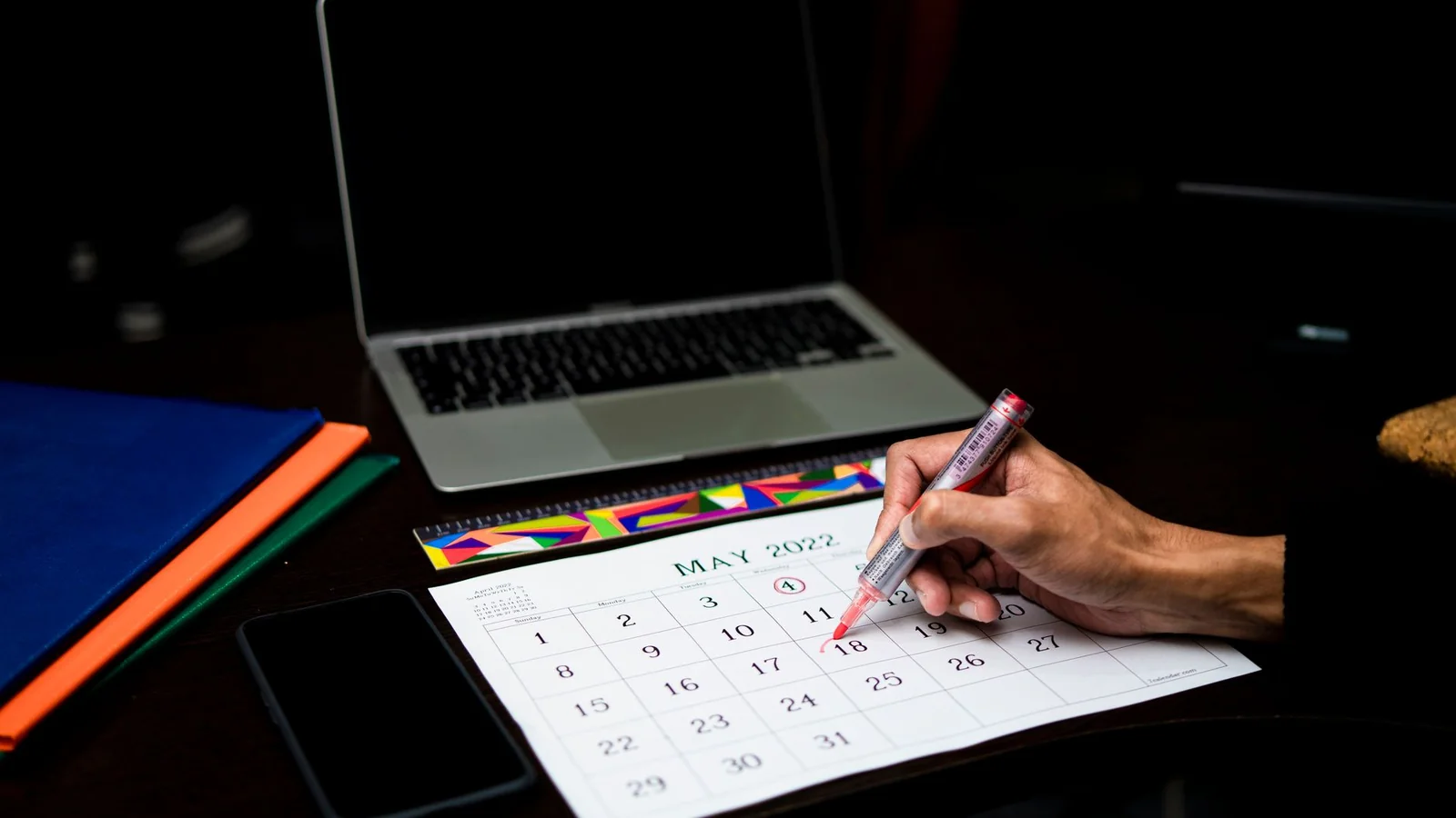 Mano marcando con un círculo una fecha en un calendario de escritorio de papel, junto a una laptop cerrada y carpetas de colores
