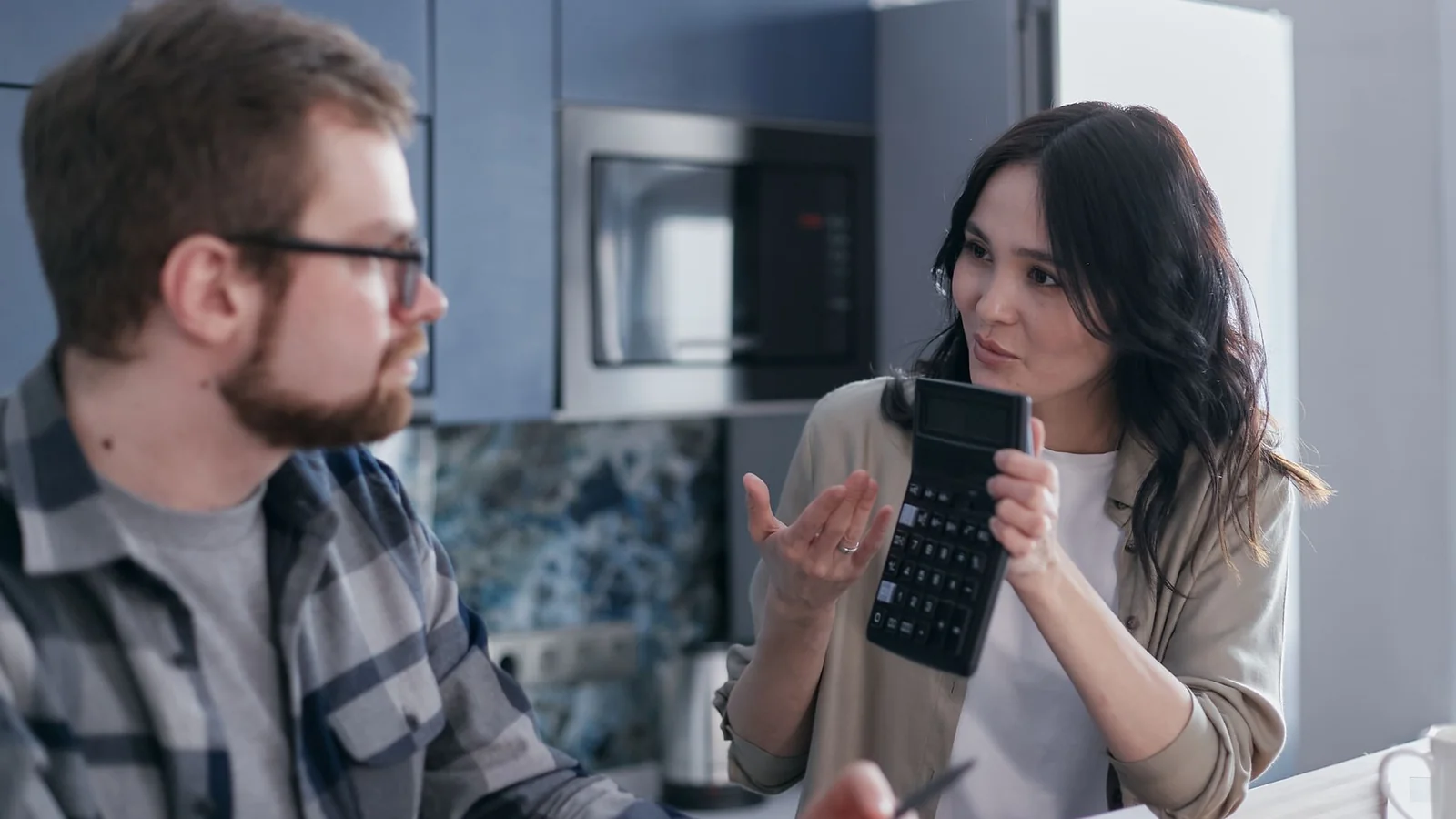 Woman holding a calculator and explaining figures to a man at a kitchen table with papers