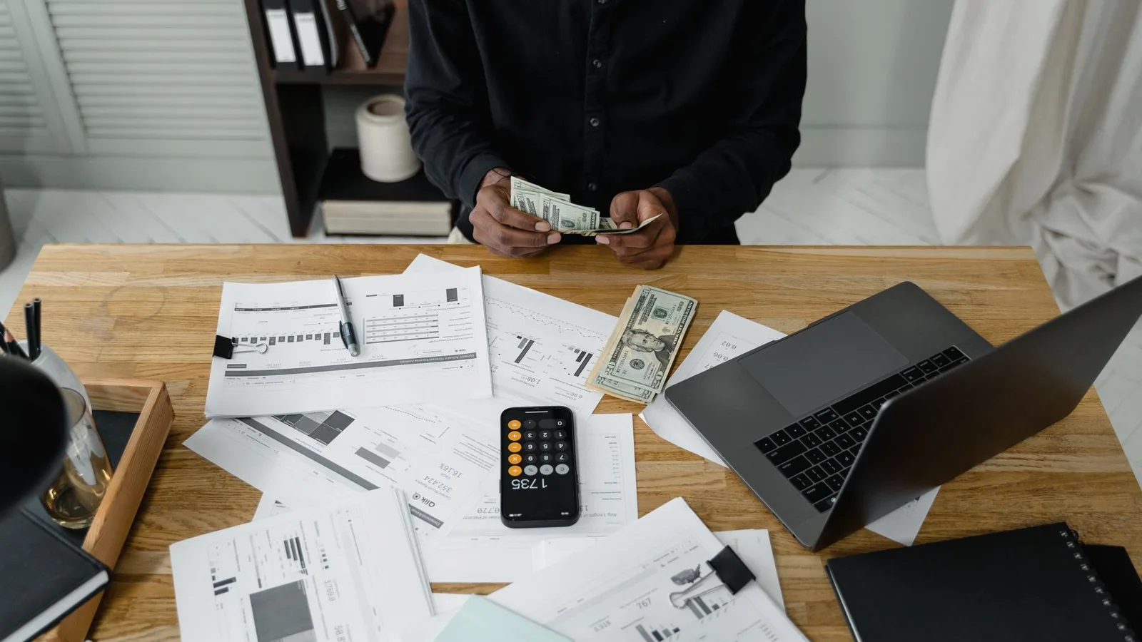 Manos contando billetes junto a registros financieros y una laptop