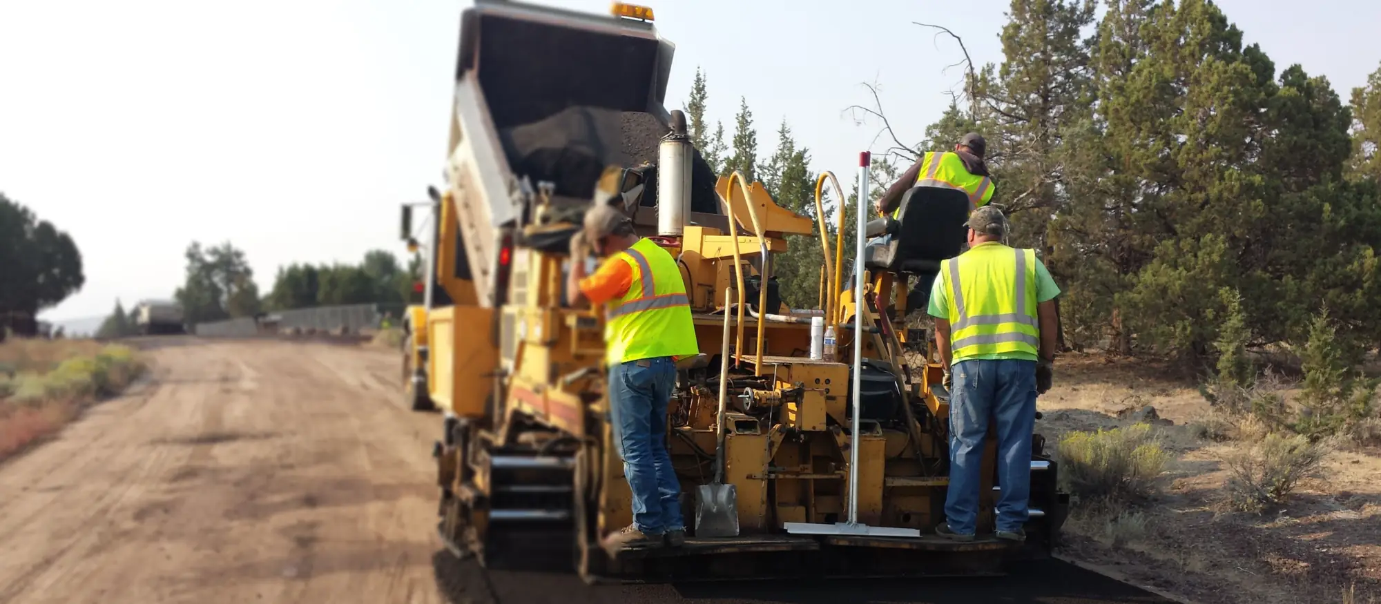 Paving crew in high-visibility vests riding an asphalt paver as it lays a fresh lane