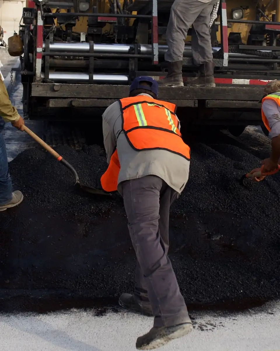Paving crew in orange safety vests shoveling hot asphalt behind a paver