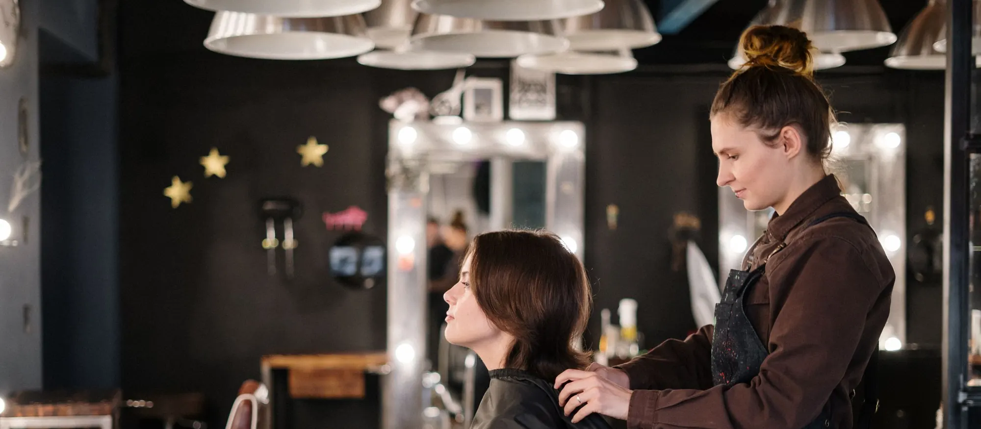 Hair stylist working on a client's cut in a salon with lighted mirrors and vintage chairs