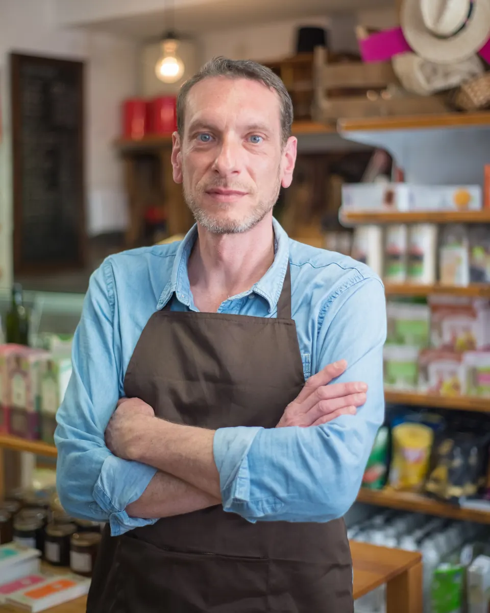 Neighborhood store owner in a brown apron and denim shirt standing with arms crossed in front of stocked shelves