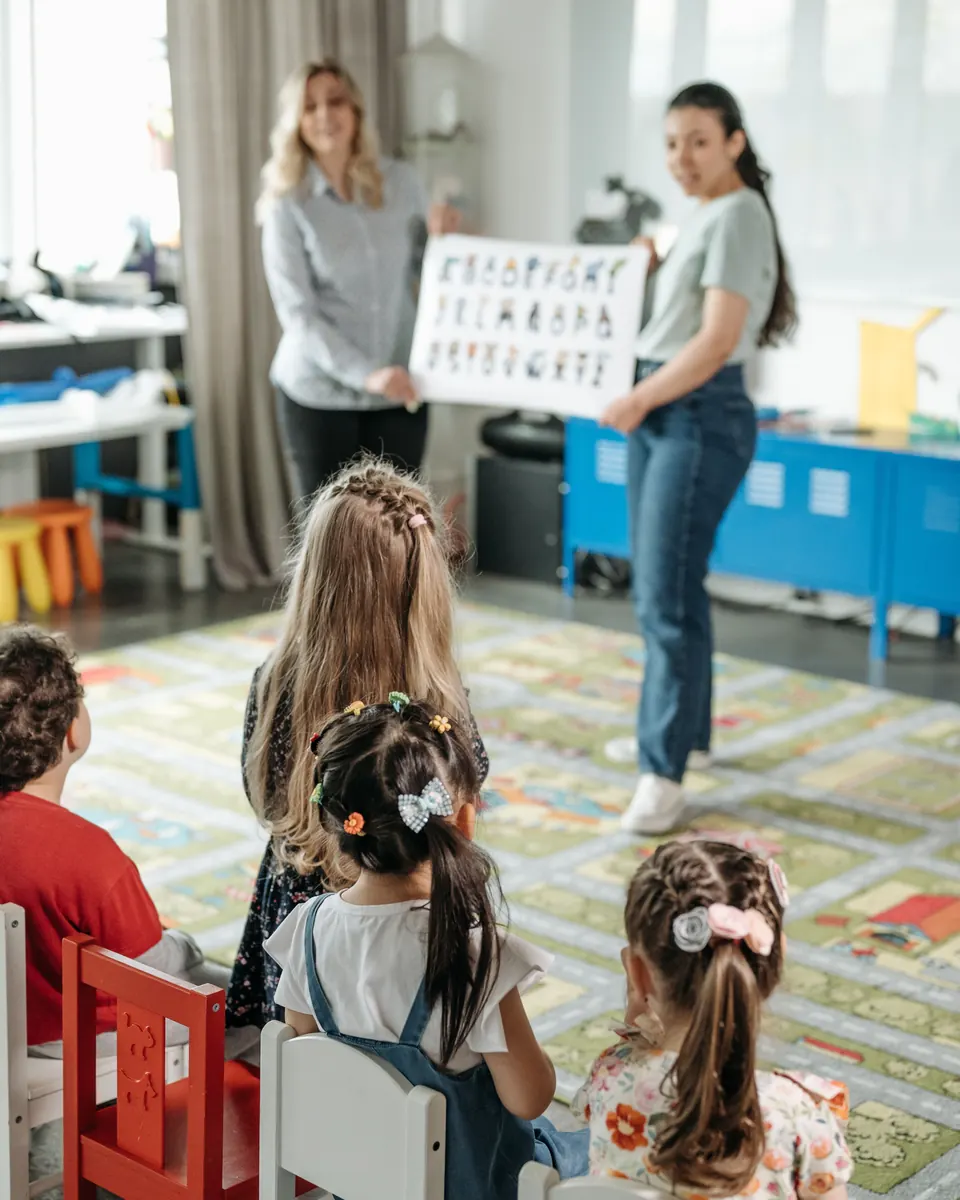 Two child care teachers holding up an alphabet picture chart for toddlers seated with their backs to the camera