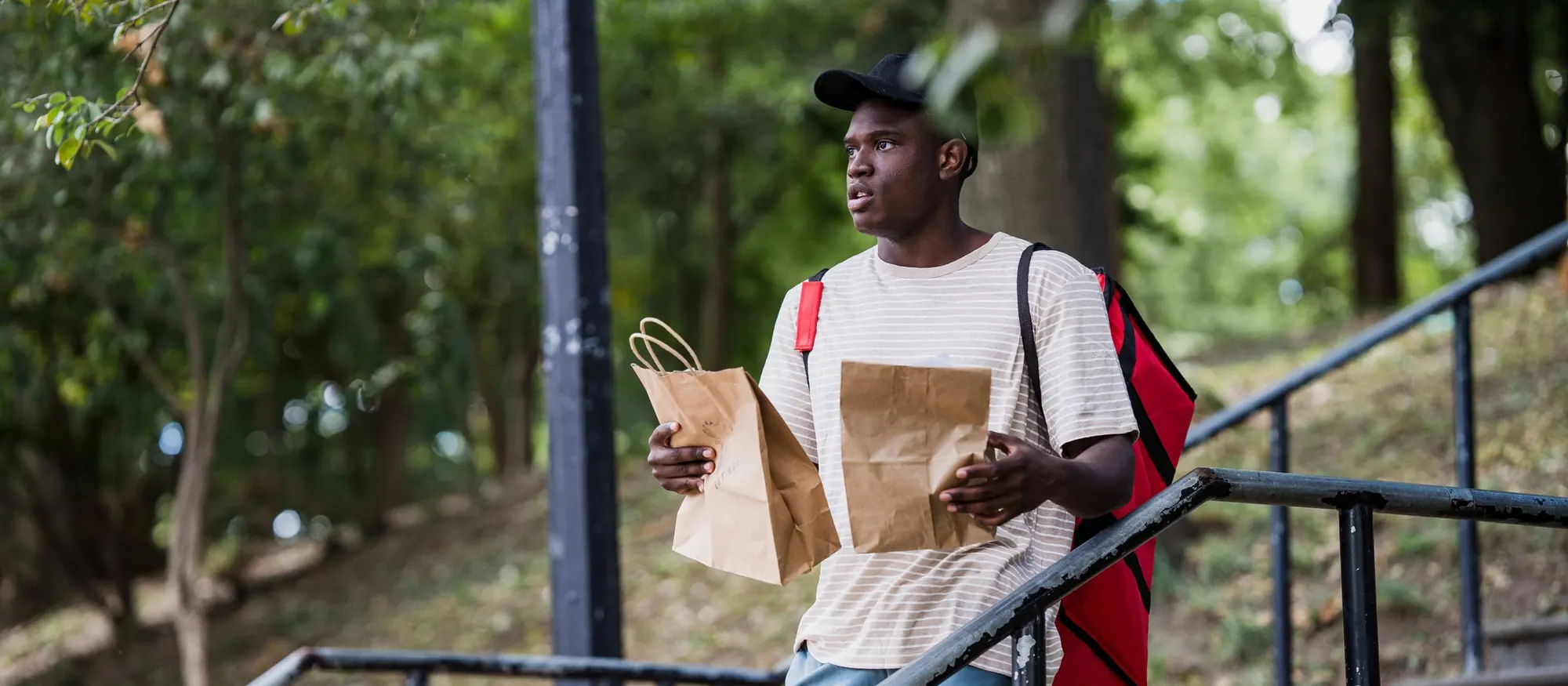 Delivery courier with an insulated backpack holding two paper takeout bags on an outdoor stairway
