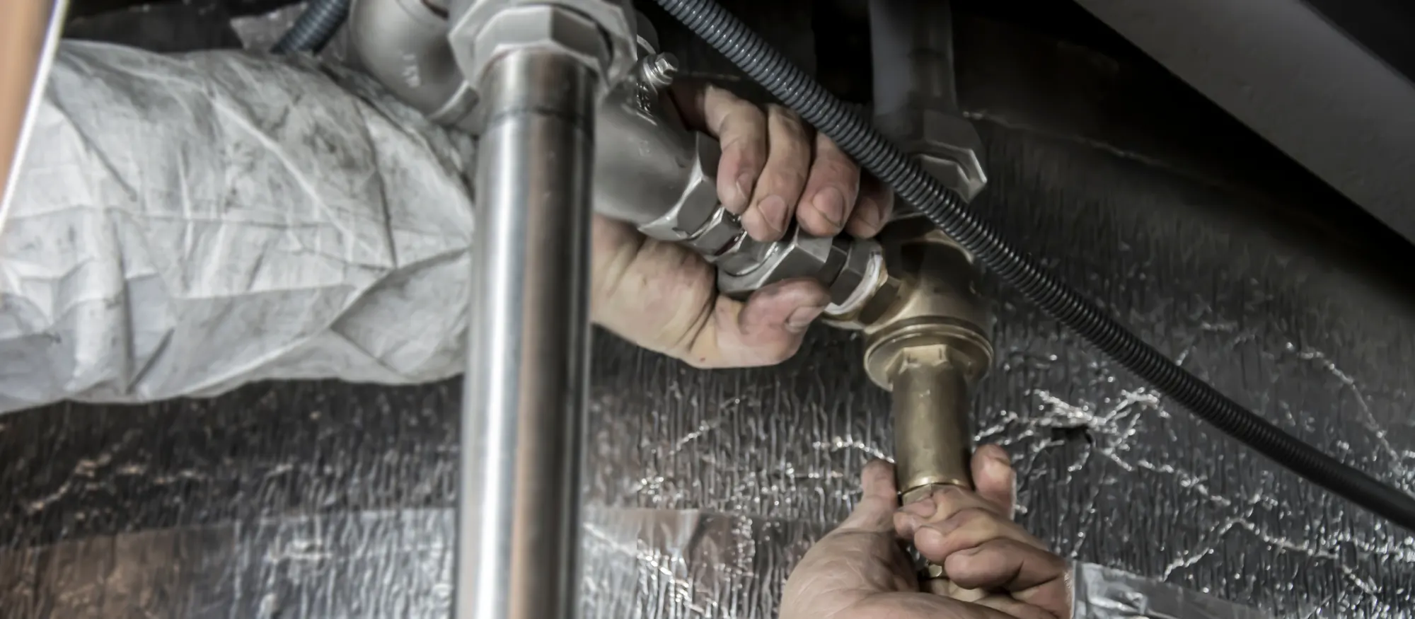 Plumber's hands threading a brass fitting onto steel water pipe beside insulated ductwork