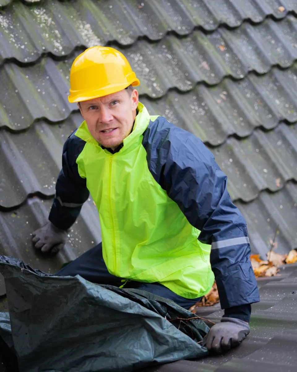 Roofer in a yellow hard hat and high-visibility jacket kneeling on a tiled roof during a repair