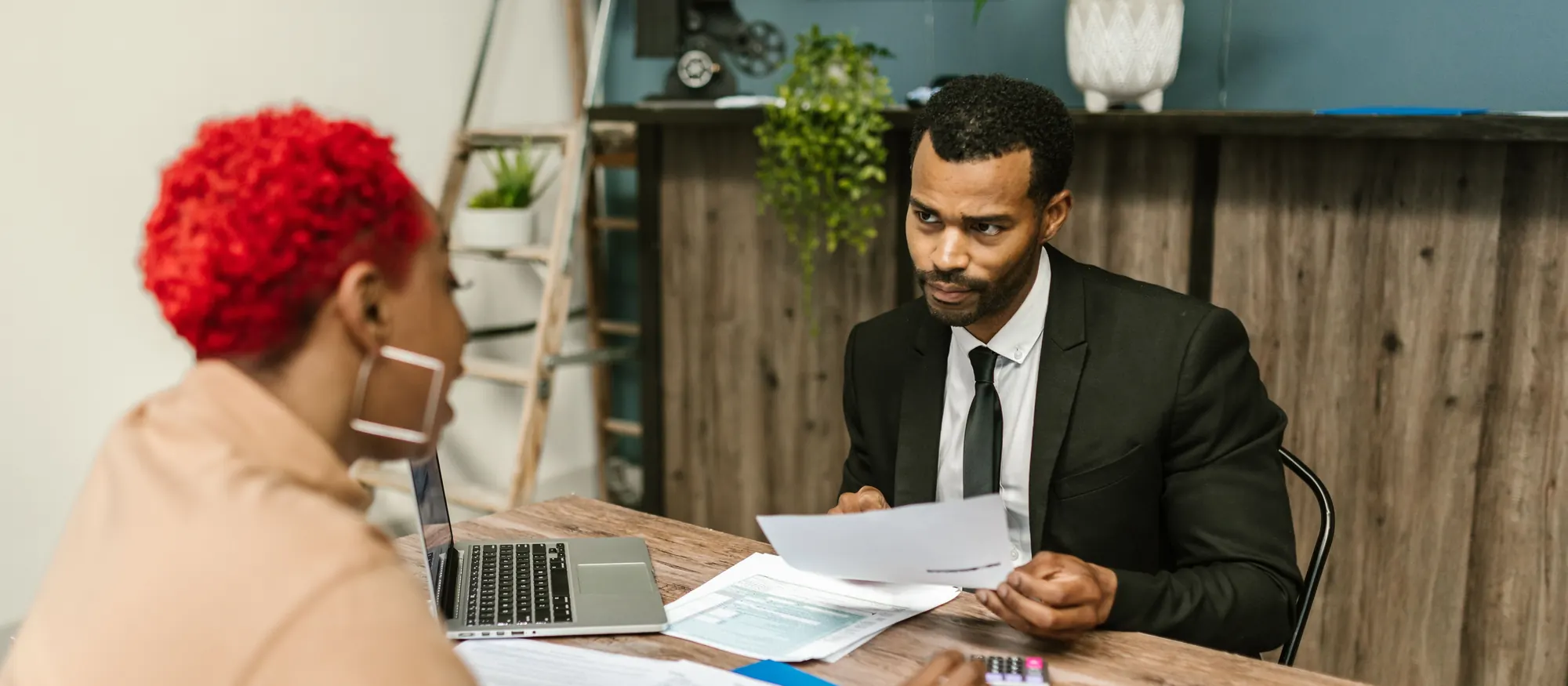 Tax preparer in a dark suit reviewing a form across a desk from a client, with a calculator and paperwork between them